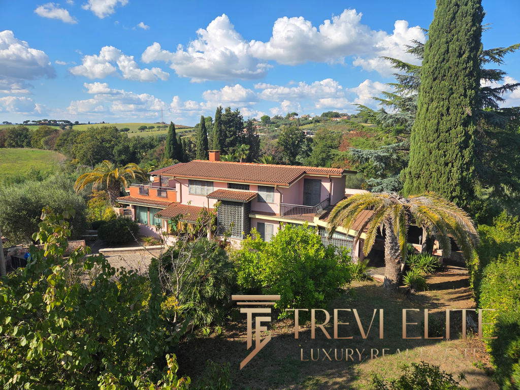 Elegant Italian villa surrounded by lush greenery and mature trees, offering a serene countryside setting with terracotta roof tiles and large windows.