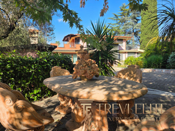 Radiant outdoor area with ornate stone table and chairs surrounded by lush greenery and mature trees, featuring a luxurious Italian residence with balconies and terracotta roof tiles in the background.