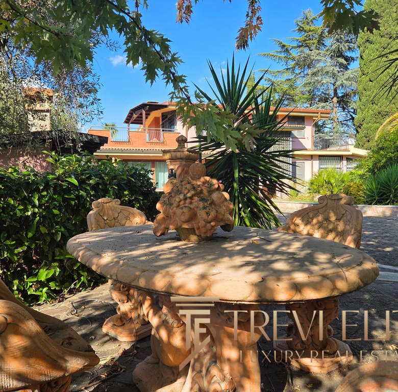 Radiant outdoor area with ornate stone table and chairs surrounded by lush greenery and mature trees, featuring a luxurious Italian residence with balconies and terracotta roof tiles in the background.