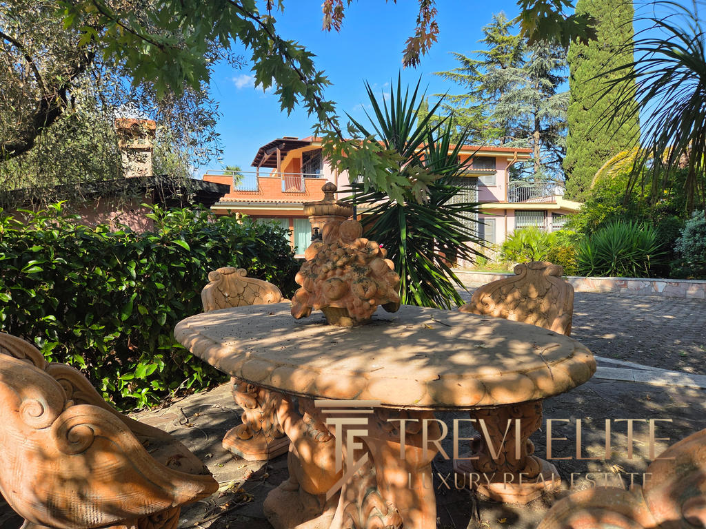 Radiant outdoor area with ornate stone table and chairs surrounded by lush greenery and mature trees, featuring a luxurious Italian residence with balconies and terracotta roof tiles in the background.