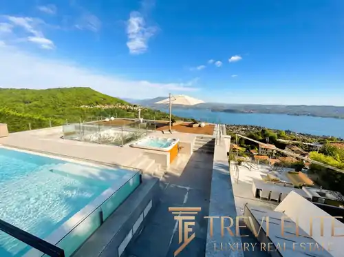 Elevated wide-angle view of a modernist luxury villa in Massino Visconti, featuring a glass-walled infinity pool and an adjacent integrated whirlpool spa. The multi-level stone terrace overlooks a 180-degree panoramic sweep of Lake Maggiore under a clear blue sky, framed by lush Mediterranean greenery and the Italian Alps in the distance.