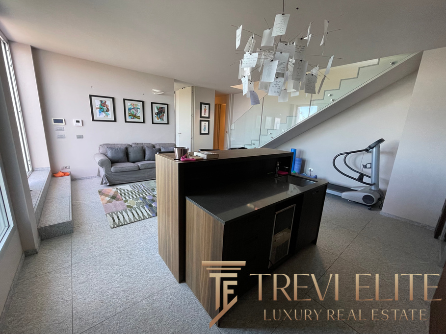 Modern living room with grey sofa, contemporary artwork, and an open-plan kitchen featuring a sleek black counter, under-stair gym equipment, and a stylish chandelier made of hanging papers.