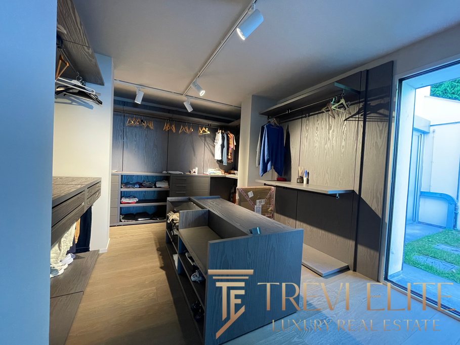 Spacious walk-in closet featuring sleek grey cabinetry, hanging space for clothes, open shelving, and vibrant natural light from a glass door opening to a lush outdoor garden.