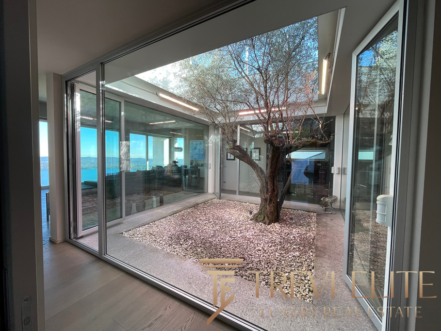 A modern indoor courtyard featuring a mature tree surrounded by white pebbles, enclosed by glass walls allowing natural light and panoramic sea views, part of the luxurious Italian real estate portfolio.