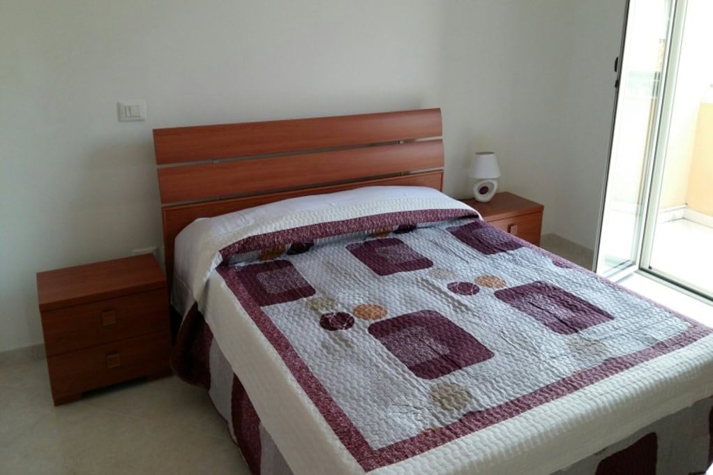 Modern bedroom with wooden furniture, double bed with colourful quilt, bedside tables, bedside lamp, and a sliding glass door leading to a balcony, in a stylish Italian property.