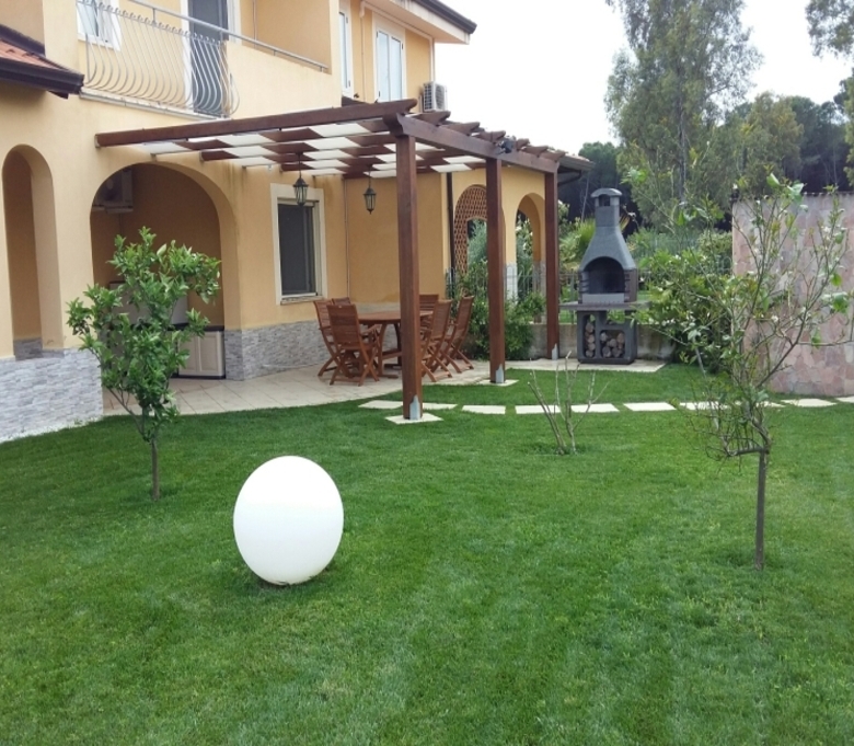 Lush green garden with young trees, a modern egg-shaped outdoor light, and a spacious patio with wooden furniture and a barbecue area at a luxurious Italian property.