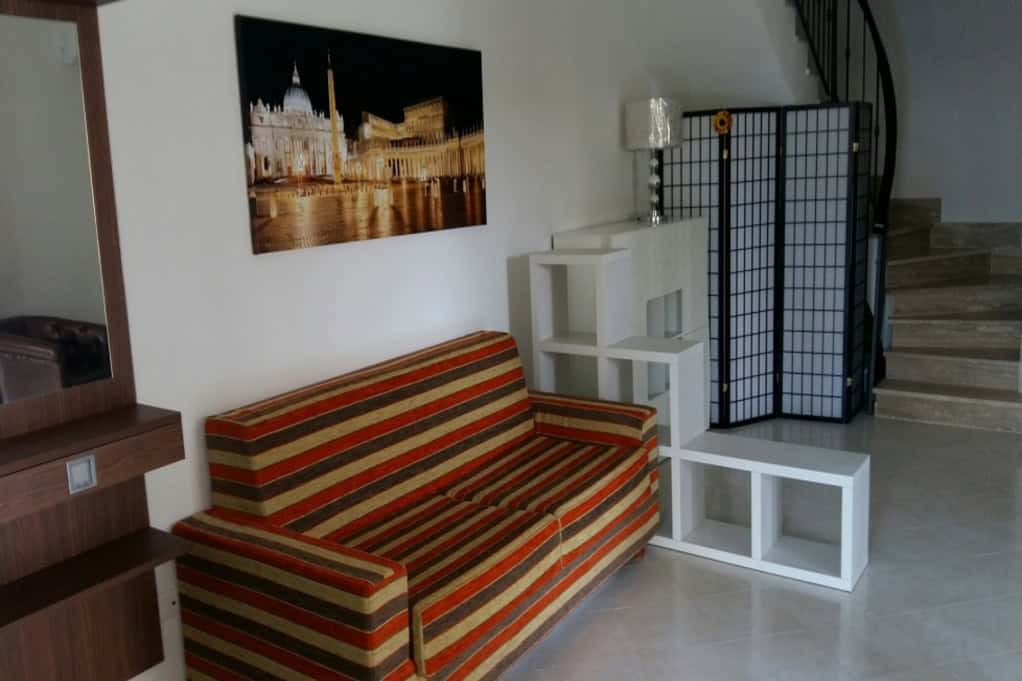 Modern living room interior with a colourful striped sofa, decorative shelving unit, artwork depicting iconic Italian landmarks, and a stylish privacy screen near a staircase.