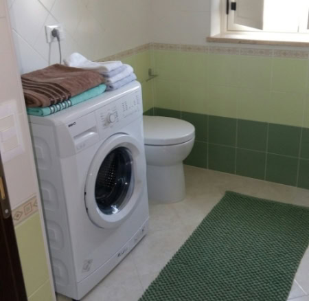 A compact bathroom with a washing machine, toilet, and natural light from a window, featuring light green and cream tiles, and a green bath mat for a fresh, clean aesthetic.