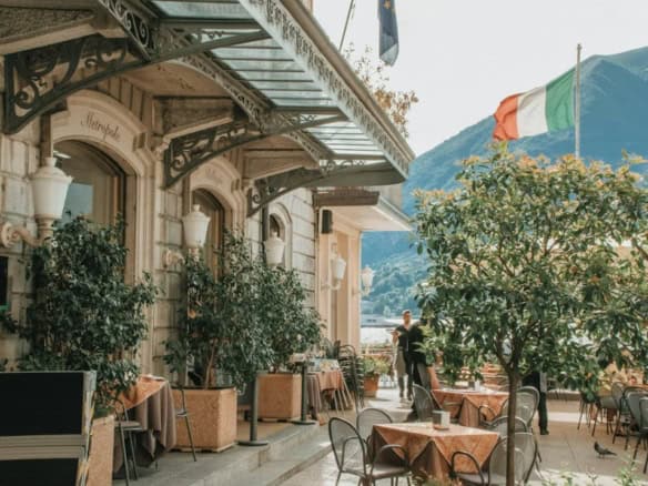 Elegant Italian restaurant terrace with lush greenery and mountain views, featuring outdoor dining with tables, chairs, and classic decor, under the Italian flag and mountain landscape.
