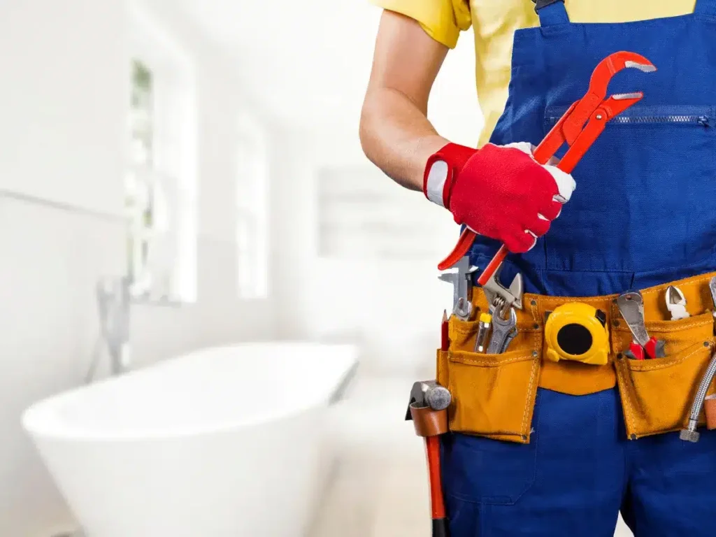 Person In Blue Overalls Holding A Red Pipe Wrench. They Are Wearing Red Gloves And A Yellow Shirt, With A Tool Belt Around Their Waist. A Bathtub Is Visible In The Background.