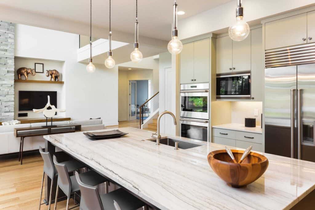 Modern Kitchen With A Large Marble Island, Hanging Pendant Lights, Stainless Steel Appliances, And A Bowl Of Utensils. A Living Area With A Sofa And Decorative Shelves Is Visible In The Background.