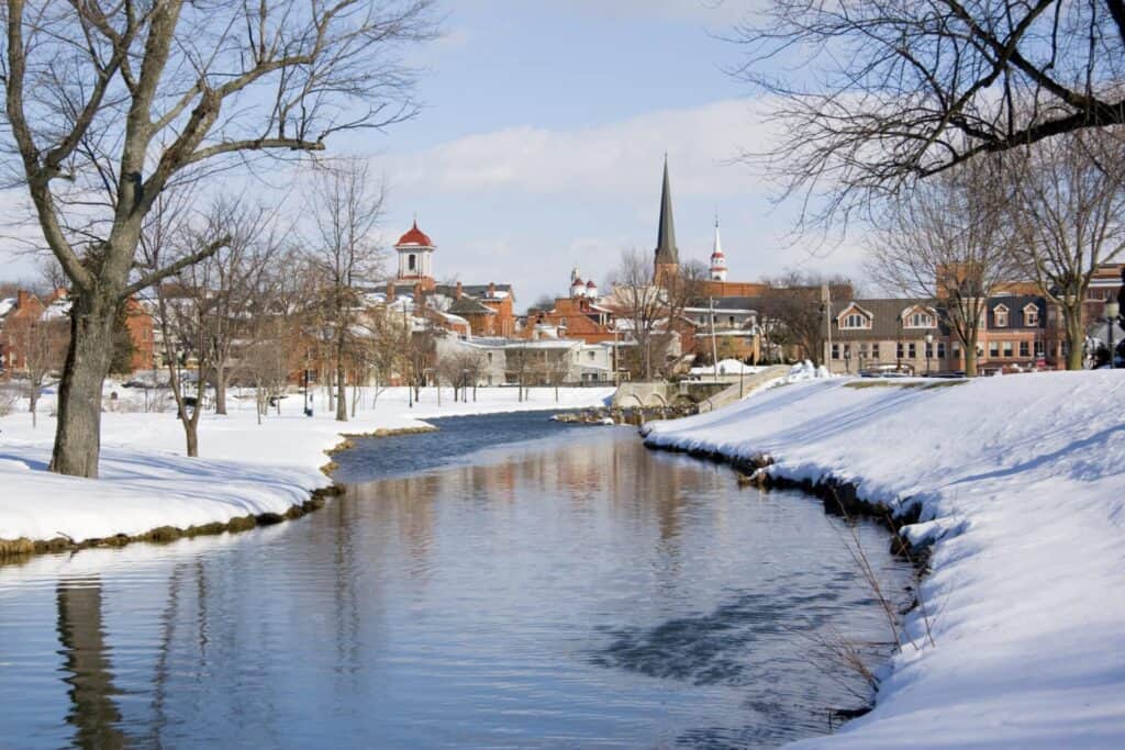 Snow-Covered Park With A River Reflecting The Sky. In The Background, A Small Town Features Historical Buildings And Church Steeples. Bare Trees Line The Riverbanks.