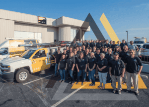 Group Of People Wearing Matching Shirts Stand In A Parking Lot Beside A Branded Company Truck With A Plumbing Logo, In Front Of A Commercial Building.