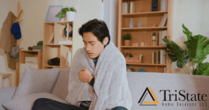 Person Sitting On A Sofa, Wrapped In A Blanket, Appearing Cold. The Room Has A Bookshelf, Coat Rack, And Decorative Items. Tristate Home Services Logo Is Visible.