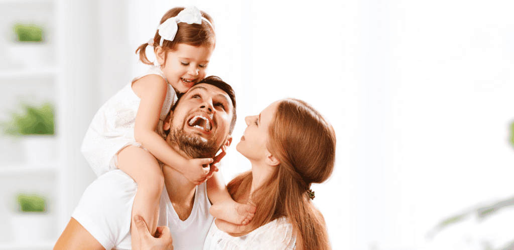 A Man Holds A Child On His Shoulders While A Woman Stands Beside Them. They Are Smiling And Appear Happy. The Background Is Bright And Blurred.