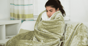 A Person Sitting On A Couch Wrapped In A Green Blanket, Appearing Cozy And Warm. Light Filtering Through A Window With Striped Curtains In The Background.