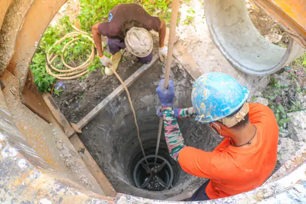 Two construction workers wearing helmets are viewed from above as they lower equipment into a deep well using a rope.