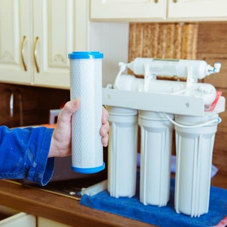 A person holds a cylindrical water filter cartridge in front of a reverse osmosis filtration system on a kitchen countertop.