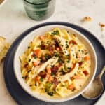 Creamy chicken and bacon pasta served with fresh bread on a black plate, garnished with chopped herbs, accompanied by green drinks, and topped with herbs and vegetables for a hearty meal.