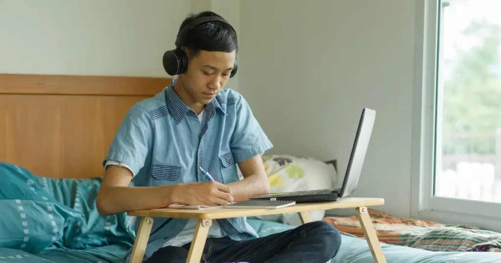 A person sitting on bed with laptop and headphones | tuitioncentre sg