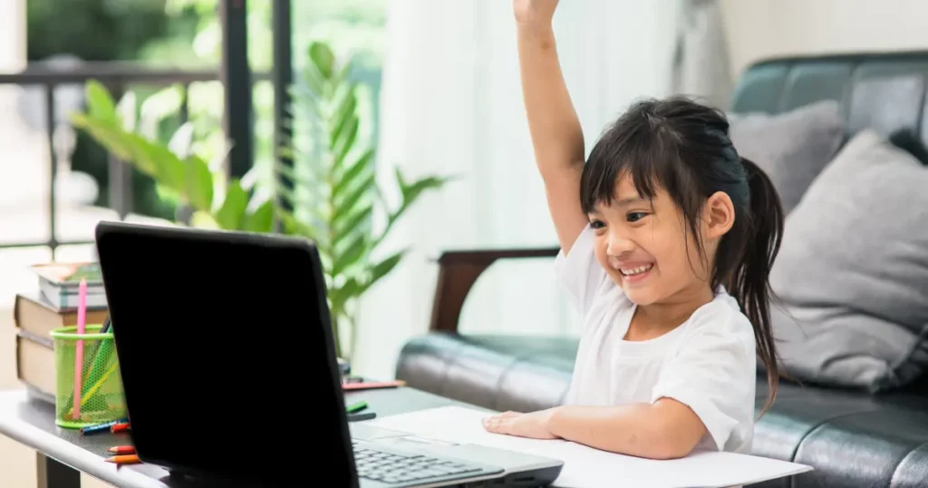 a child is sitting in front of a laptop and raising their hand | tuitioncentre sg