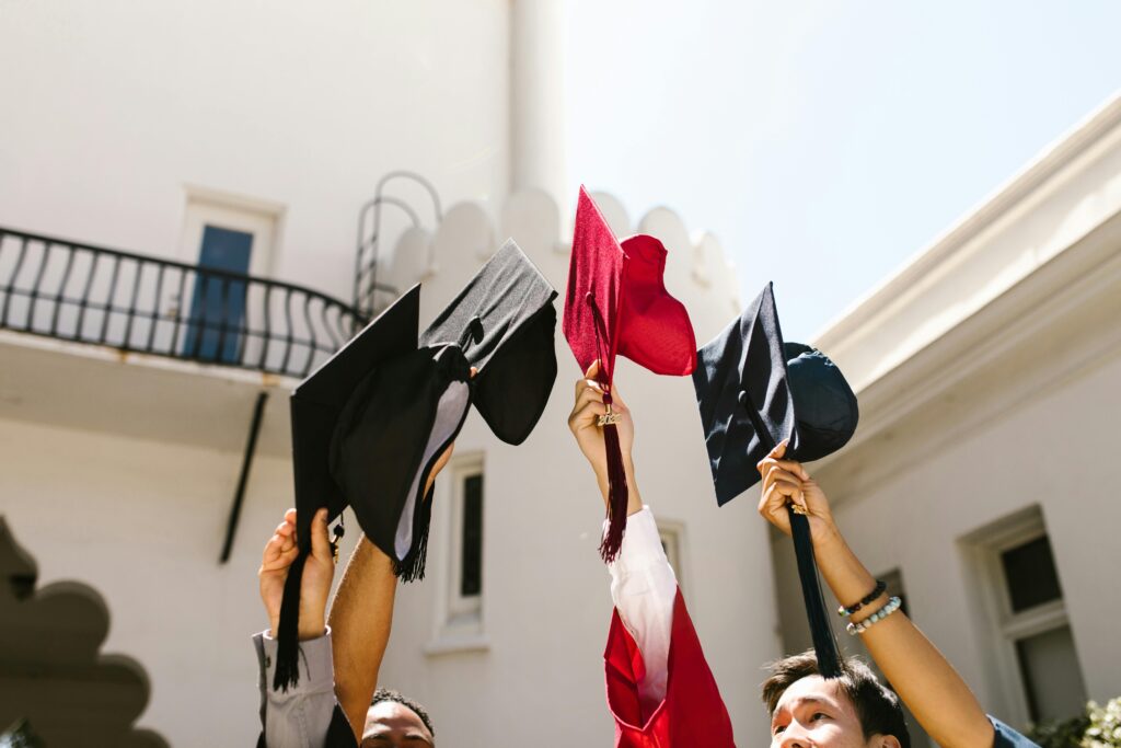 Graduates celebrate, holding up black and red caps against a bright sky and white building. A moment of triumph and new beginnings | Tuition Centre SG