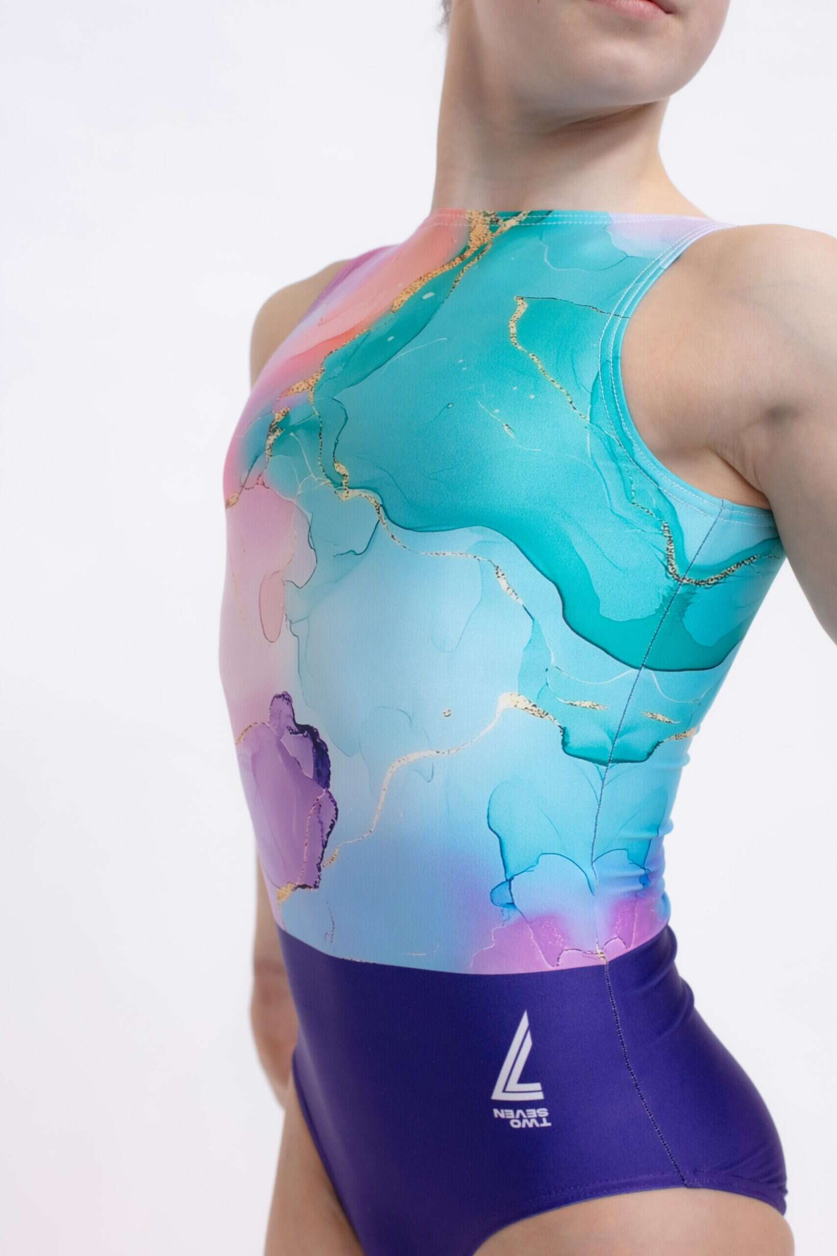Primer plano del torso delantero de una gimnasta joven con justaucorps marmolado en tonos turquesa, malva y rosa con sutiles vetas doradas, mostrando el logotipo de la marca en la cadera, sobre fondo blanco.
