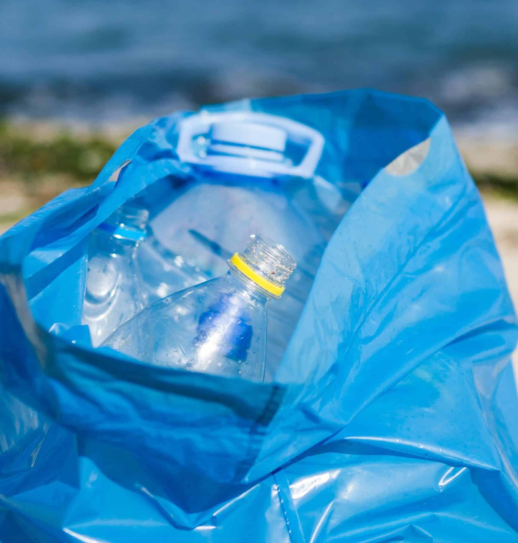 FOTO SOSTENIBILIDAD Bolsa azul de reciclaje llena de botellas de plástico transparentes en primer plano, con un fondo desenfocado que muestra el mar.