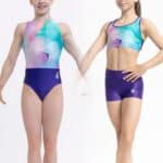 Dos gimnastas jóvenes de pie sobre fondo blanco, una vistiendo un justaucorps marmolado en tonos turquesa, malva y rosa y la otra un conjunto de top deportivo y shorts a juego; ambas posan de frente mostrando el diseño y el logotipo de la marca.