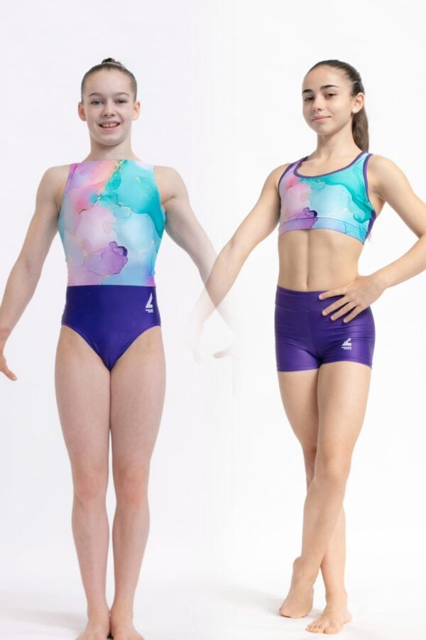 Dos gimnastas jóvenes de pie sobre fondo blanco, una vistiendo un justaucorps marmolado en tonos turquesa, malva y rosa y la otra un conjunto de top deportivo y shorts a juego; ambas posan de frente mostrando el diseño y el logotipo de la marca.