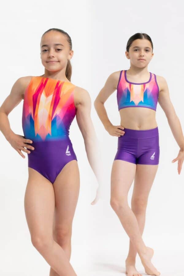 Dos gimnastas jóvenes posando de frente sobre fondo blanco; a la izquierda llevan un justaucorps marmolado en tonos turquesa, malva y rosa, y a la derecha un conjunto de top deportivo y shorts a juego en los mismos colores, ambas en posición de baile mostrando el diseño y el logotipo de la marca.