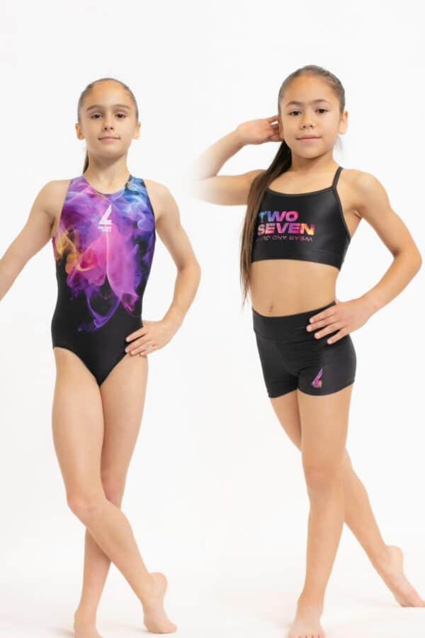 Dos gimnastas jóvenes posando de frente sobre fondo blanco; la de la izquierda viste un justaucorps negro con estampado de humo en colores vibrantes (rosa, púrpura, amarillo y azul), y la de la derecha un conjunto negro de top deportivo con texto “TWO SEVEN” en degradado y shorts a juego, ambas en posición de baile mostrando el logotipo de la marca.