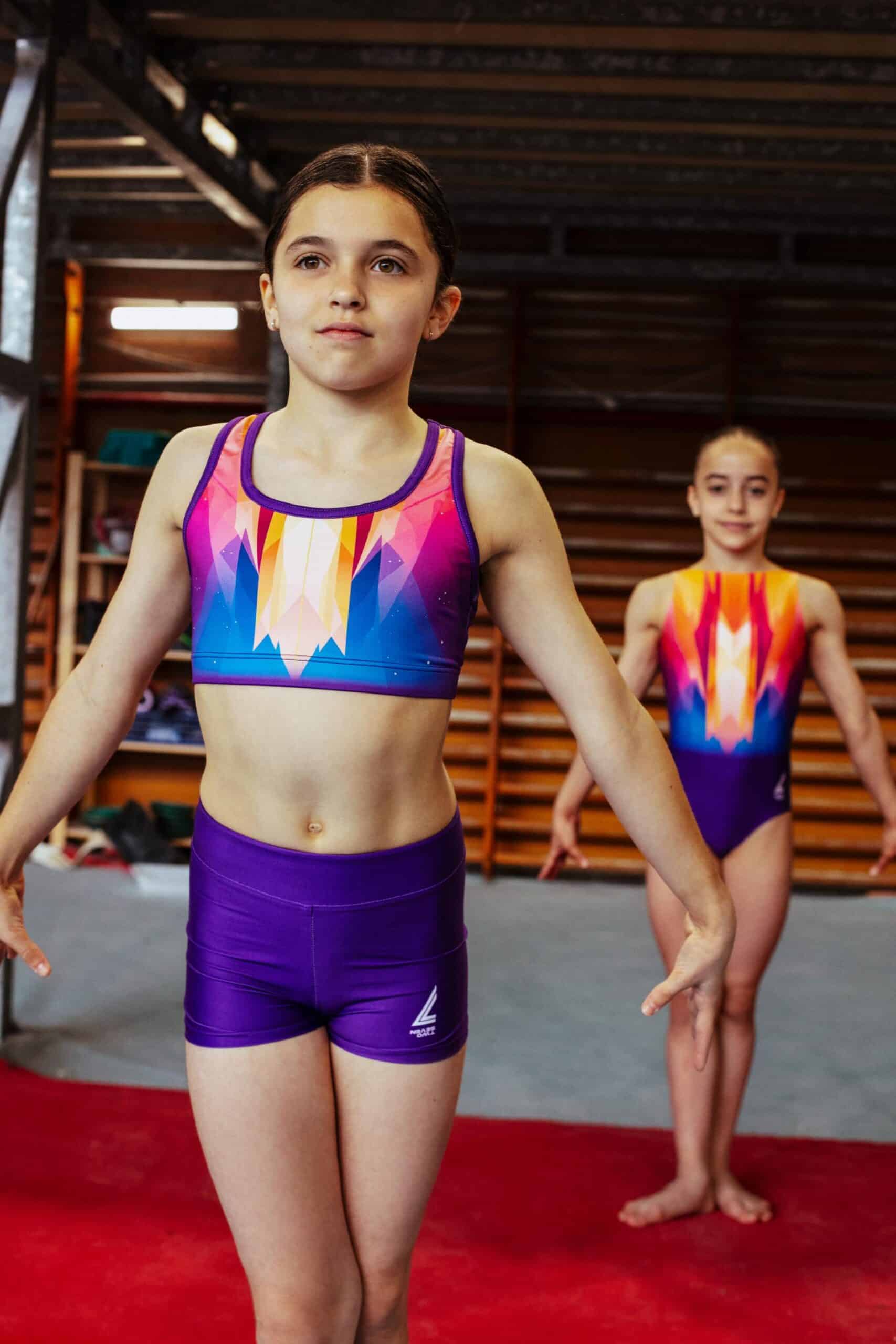 Dos gimnastas en el gimnasio: una en primer plano de pie y otra al fondo, ambas vistiendo el conjunto Prisma Glow con top y shorts