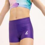 Detalle frontal de una gimnasta joven vistiendo un top deportivo marmolado en tonos turquesa, malva y rosa, combinado con shorts ajustados morados con el logotipo de la marca, sobre fondo blanco.
