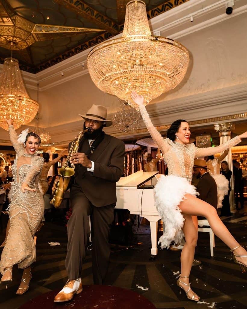 Elegant dancers and saxophonist performing at a luxurious event with grand chandeliers and classic decor.