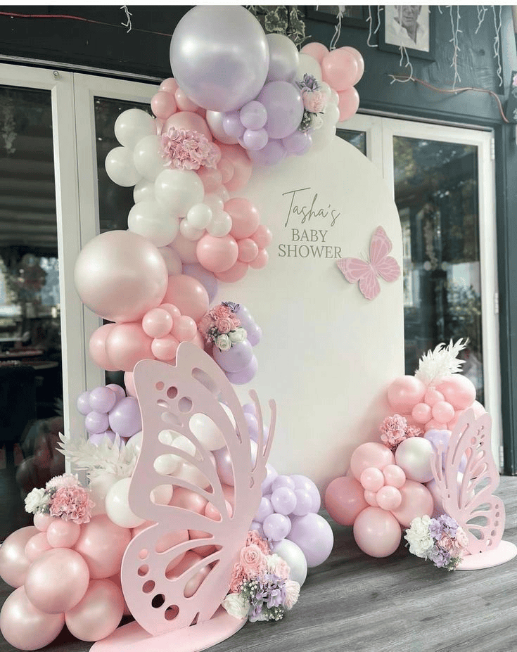 Pink and purple balloon birthday shower backdrop with floral arrangements and butterfly decorations.