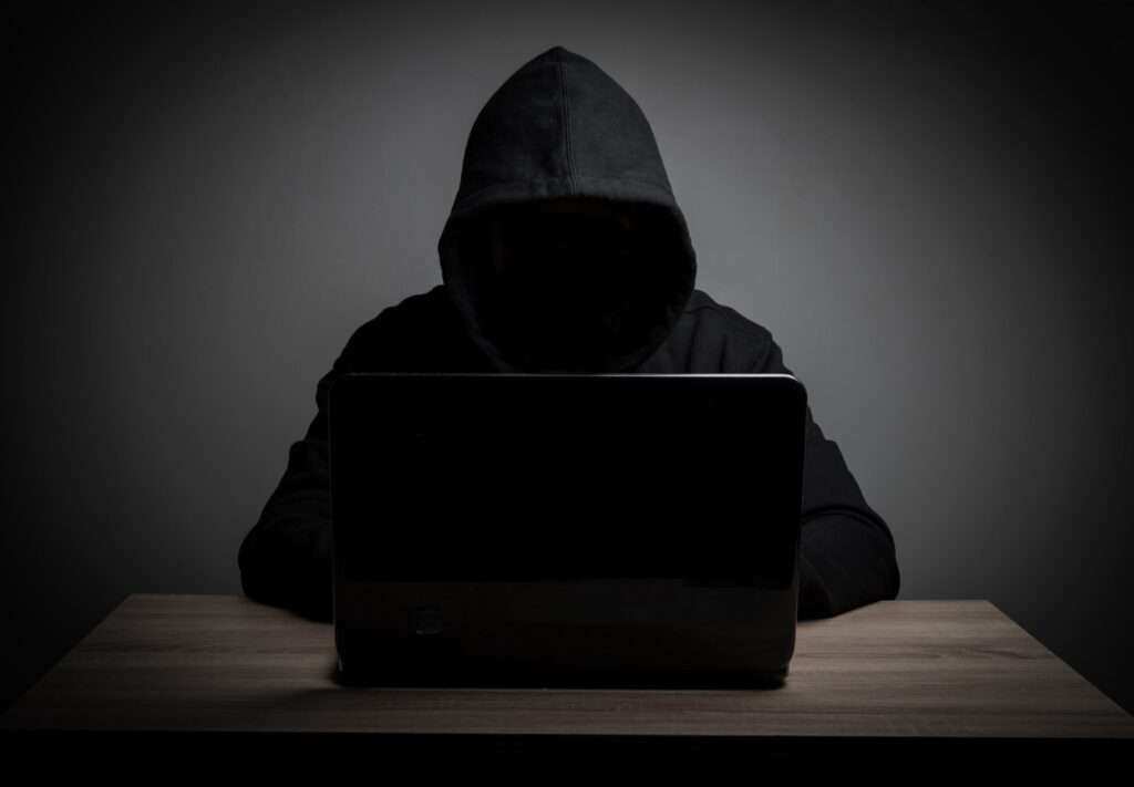 Sötétben ülő, kapucnis hálózati bűnöző laptop előtt.
