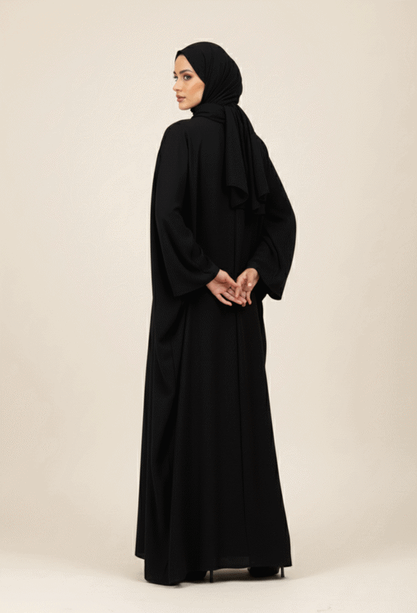 Elegant woman in black abaya and hijab standing against a neutral background, showcasing modest fash.