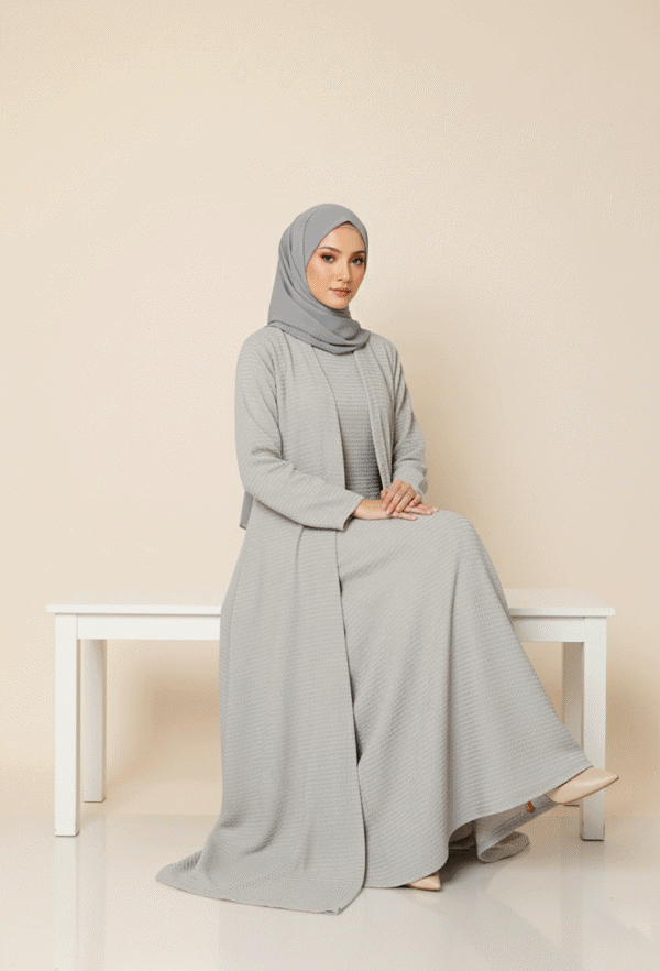 A woman wearing a modest, elegant grey abaya and hijab, sitting on a white bench against a neutral b.