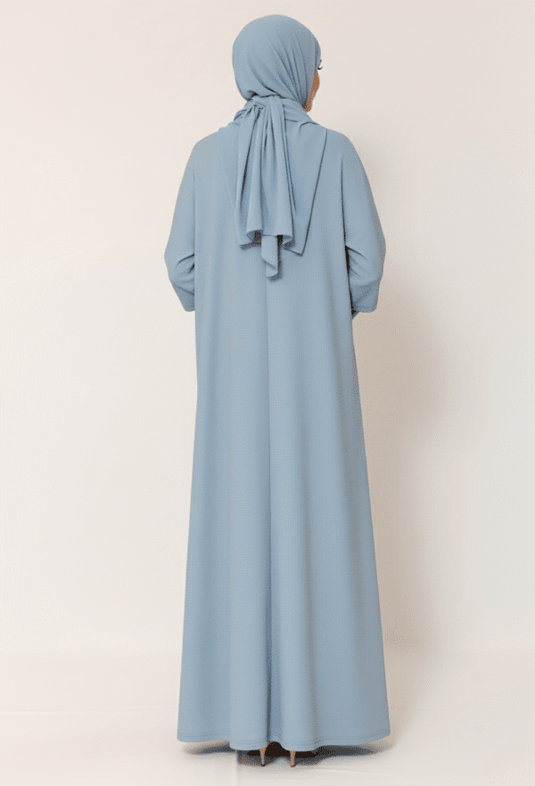 Elegant woman in light blue modest abaya and hijab, standing against a neutral background.