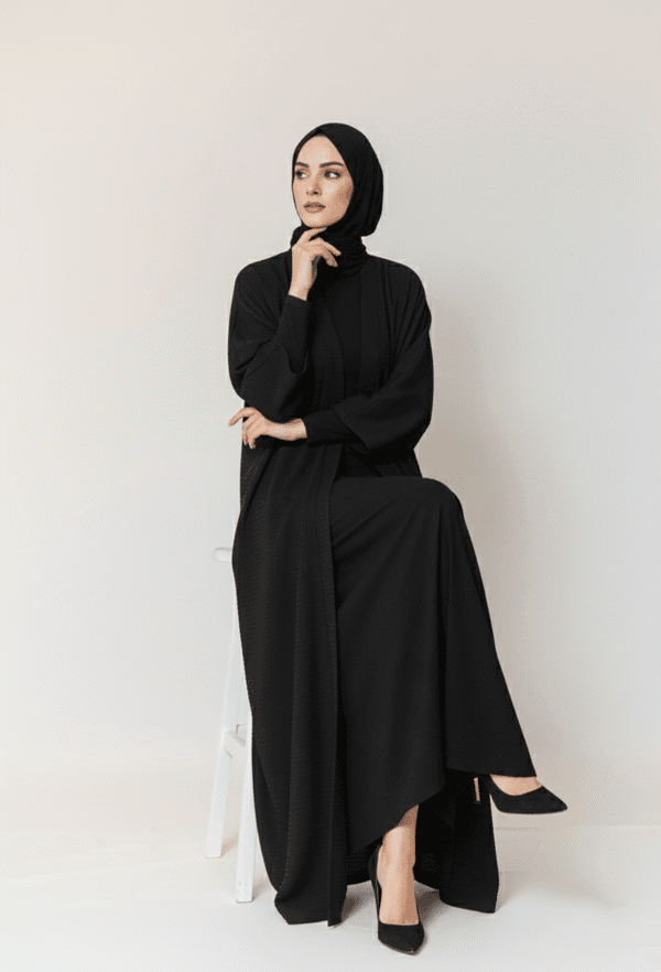 Woman in elegant black modest dress and hijab, sitting on a white stool against a plain background.