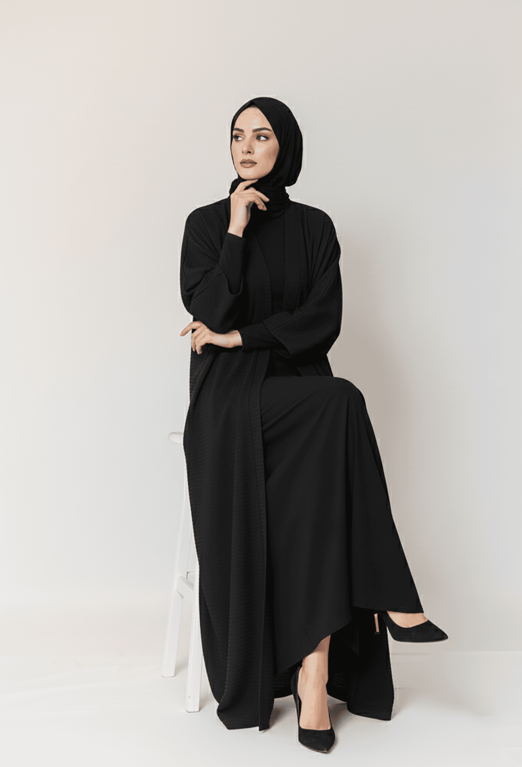 Woman in elegant black modest dress and hijab, sitting on a white stool against a plain background.
