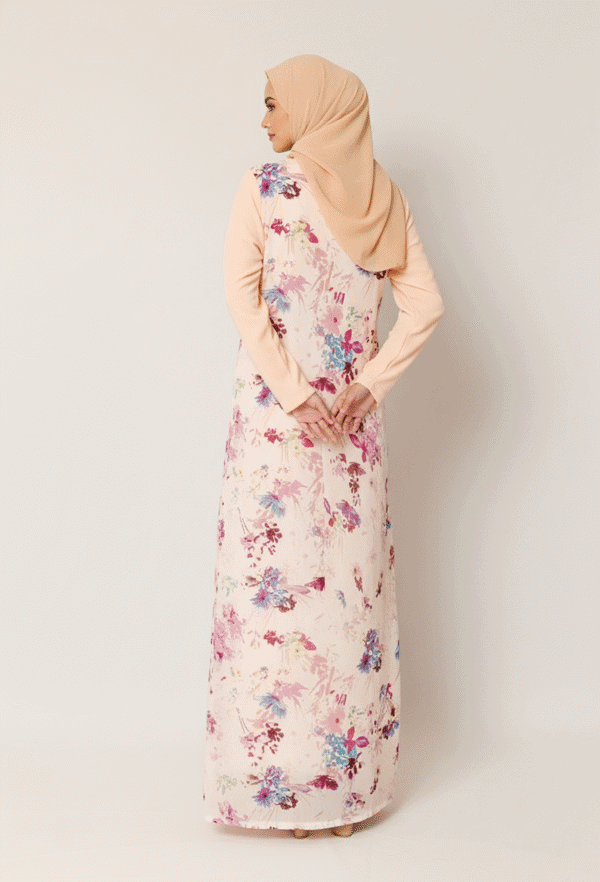 Elegant woman in floral modest dress with beige hijab, standing against a plain background.