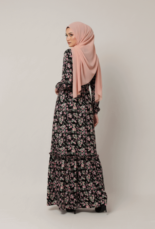 A woman wearing a floral maxi dress and a pink hijab, standing against a plain background.