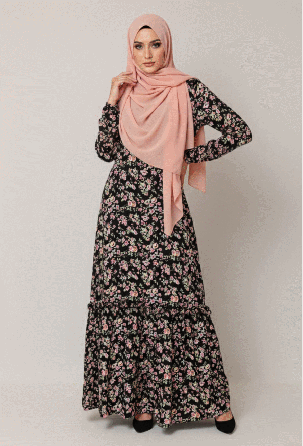A woman wearing a floral maxi dress and a pink hijab, standing confidently against a plain backgroun.