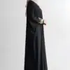 Woman wearing a black abaya with gold embroidery on sleeves, standing sideways.
