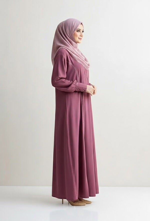 Woman in pink abaya and hijab standing sideways against a plain background.