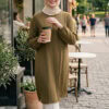 A smiling woman in a beige hijab and olive green dress holding coffee outside a café in a lively cit.