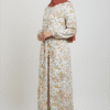 Woman in floral maxi dress and hijab, smiling, standing against a plain background.