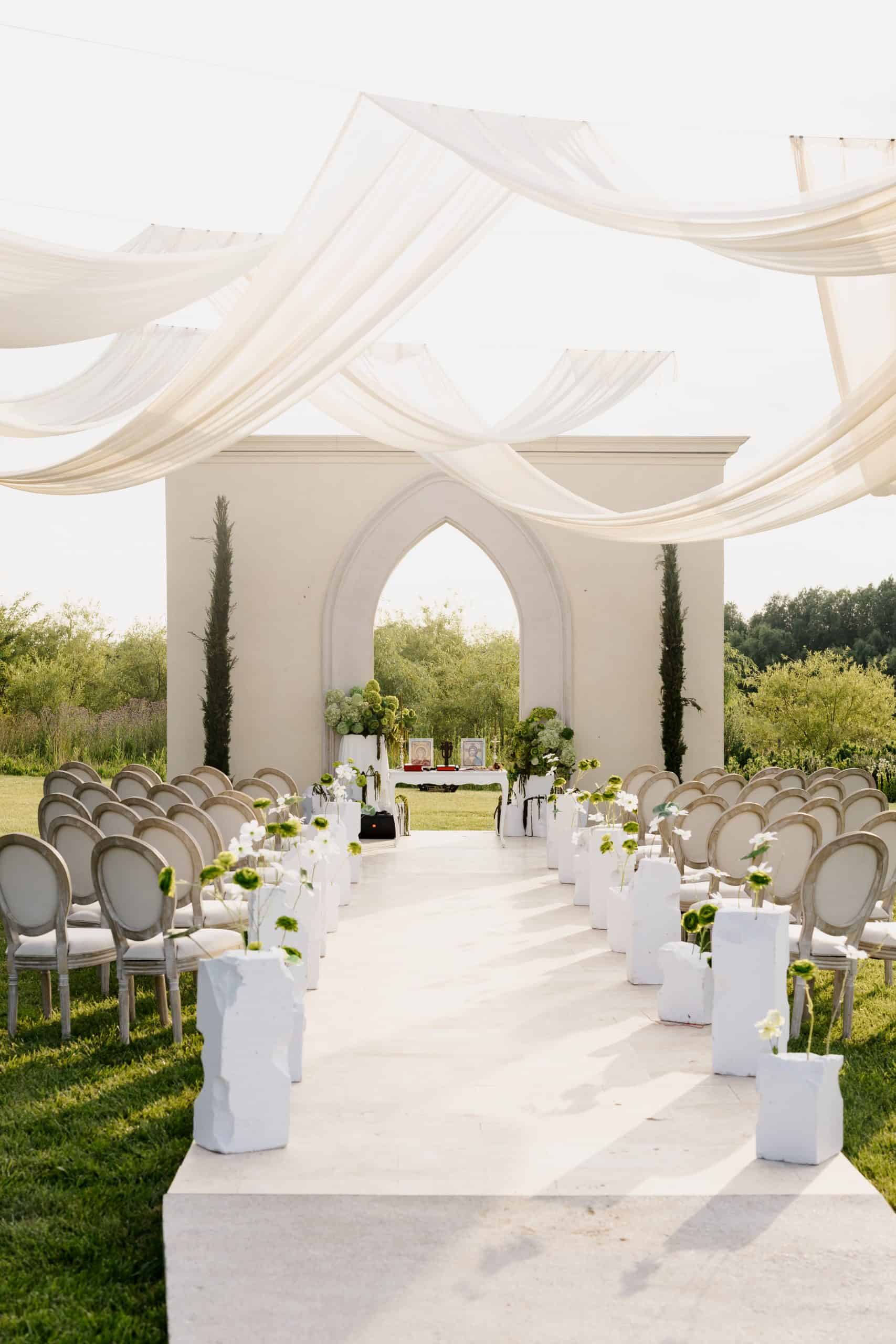Elegant outdoor wedding ceremony setup featuring white and green floral arrangements, draped fabric, and a scenic natural background.
